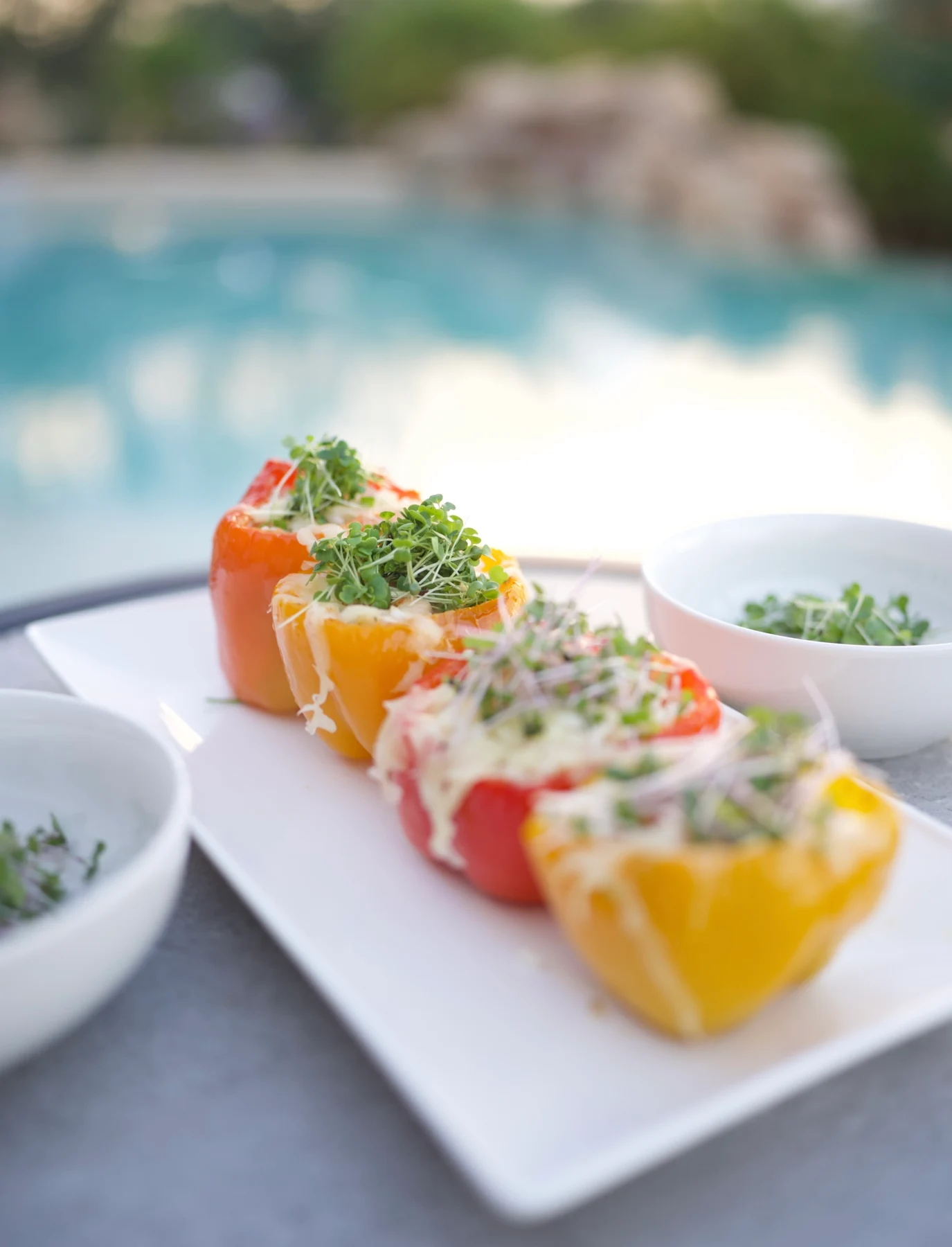 Red and yellow stuffed peppers topped with fresh microgreens on a white platter beside the pool